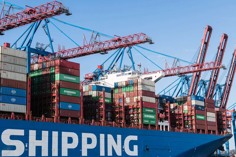 Stacked shipping containers being loaded onto a vessel by port gantry cranes